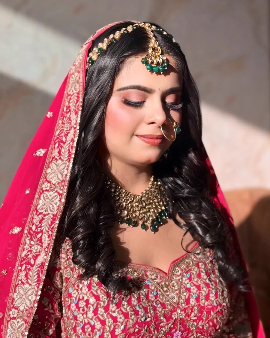 One of our brides in a deep pink lehenga with green and gold kundan jewellery, eyes lowered