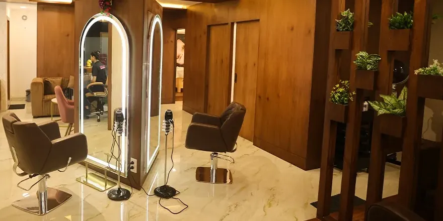 Two styling chairs facing lit arch mirrors on a marble floor, next to a wooden slat divider planted with greenery.