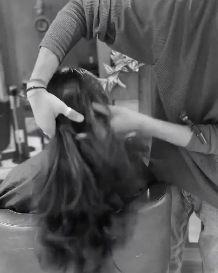 Black-and-white view from behind of a stylist's hand lifting long dark hair at the salon chair.