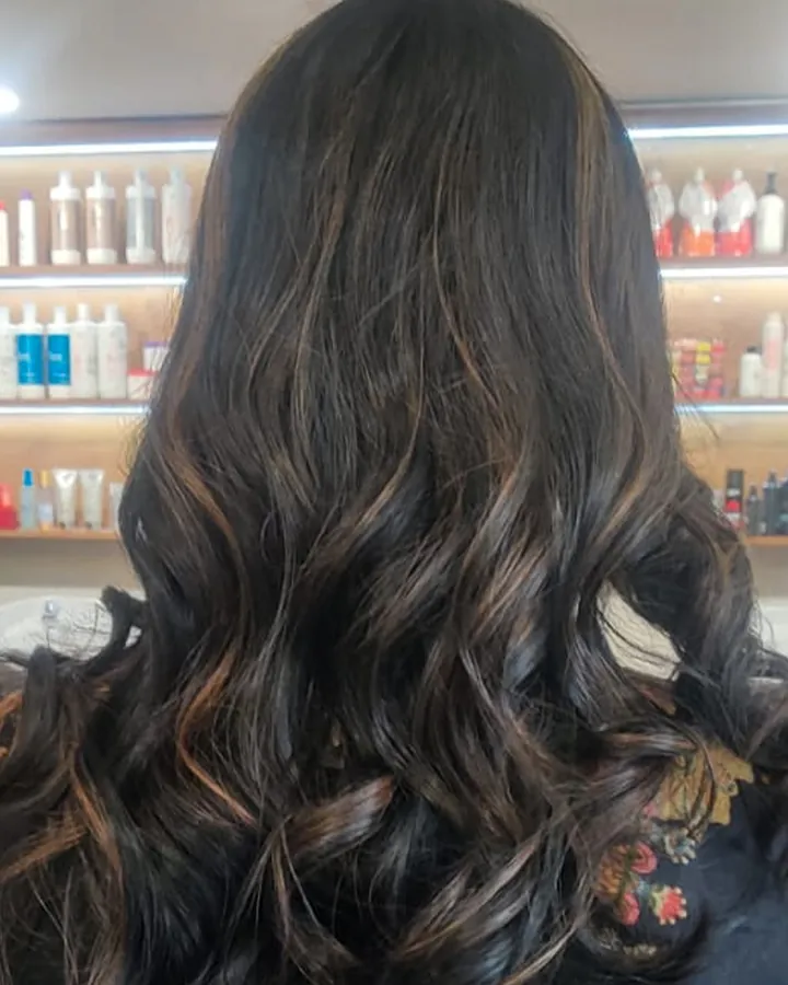 The back of a seated client's head, long dark hair with warm caramel-brown ribbons through it.