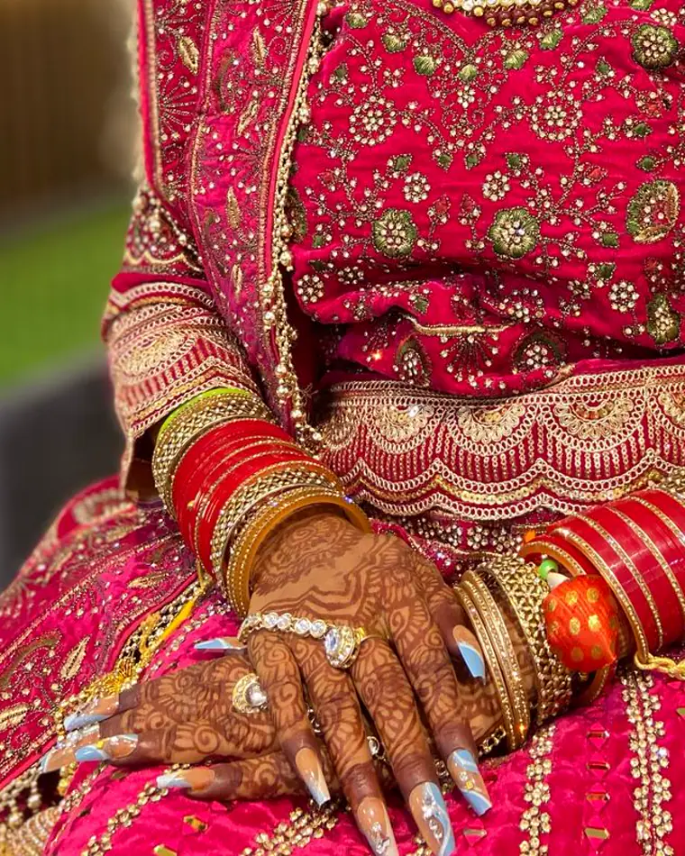 A bride's hands with mehndi, red chooda and long pale blue marbled nail extensions.