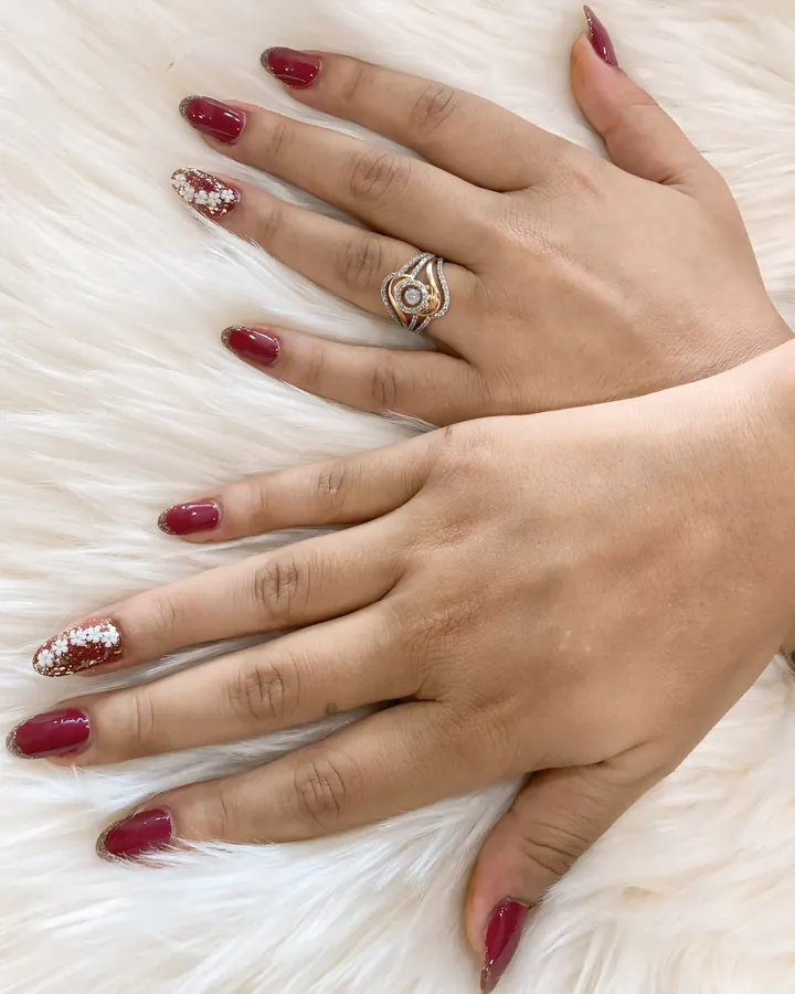 Two hands resting on a white fur throw with maroon almond nails and two accent nails in white flowers and gold glitter.