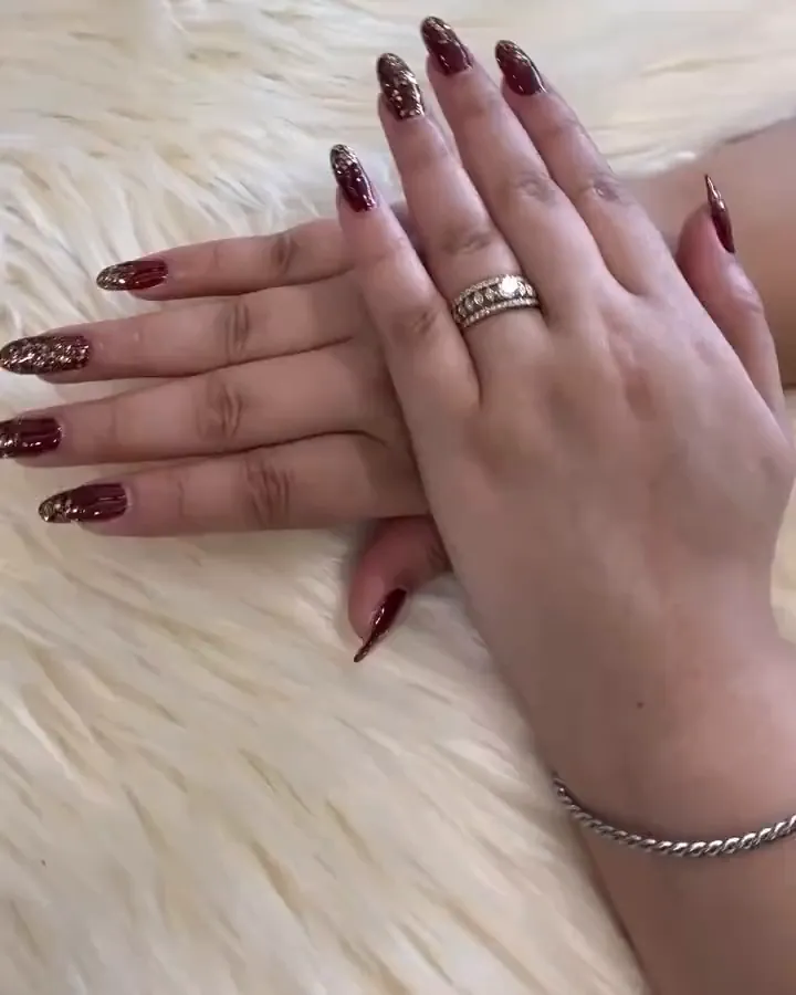 Two hands on a white fur throw with dark maroon almond nails and gold glitter accent nails.