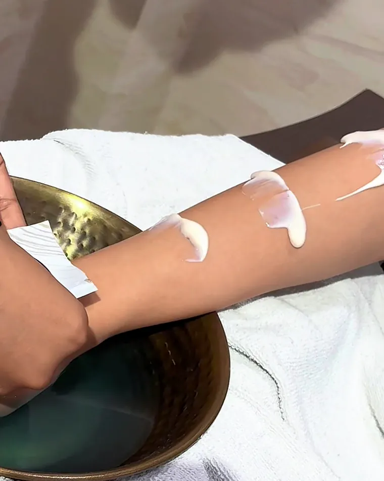 A forearm with cream on it beside a brass bowl of water, on a folded white towel.