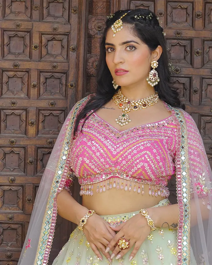 Haldi look: a hot-pink mirror-work blouse with a mint lehenga and a navratna necklace at a carved door.