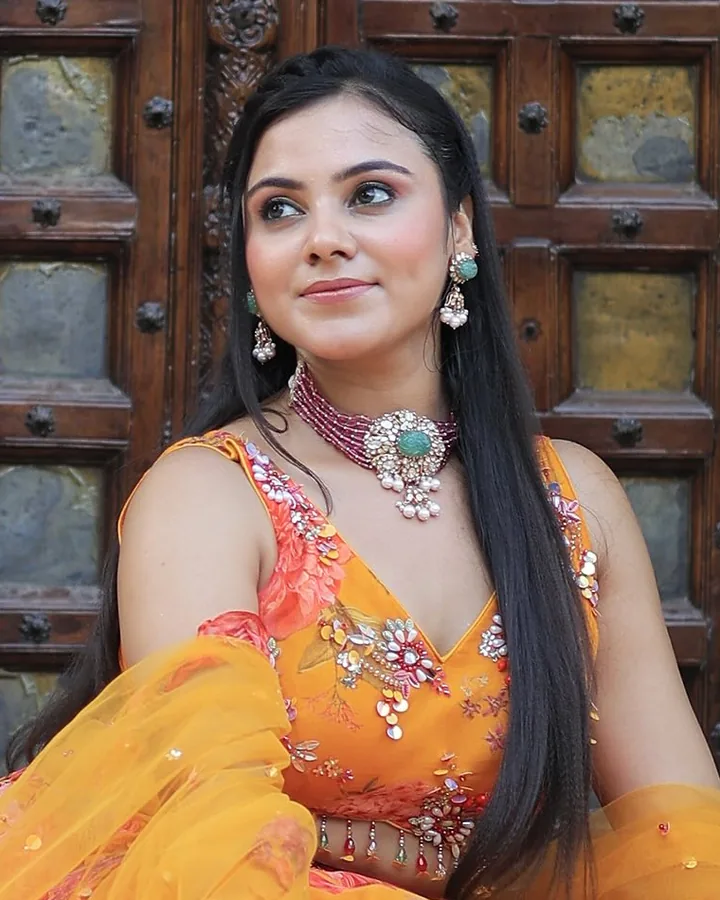 Haldi look: an orange floral outfit with a red bead choker, seated at a carved door, from our styled shoot.