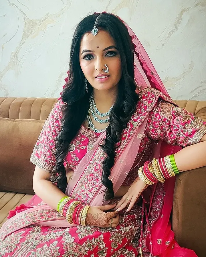 A bride in a pink and magenta embroidered lehenga sits on a beige sofa with her veil draped over her head and stacked bangles on both wrists.
