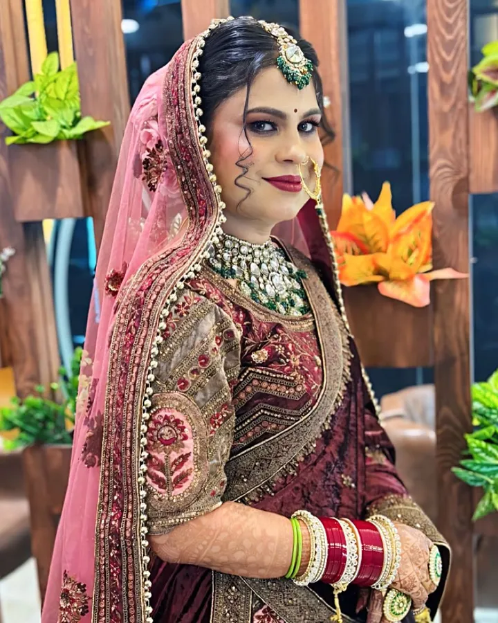 A bride in a maroon velvet lehenga with a pink veil stands beside a wooden partition, hands folded, wearing a green and white kundan choker.