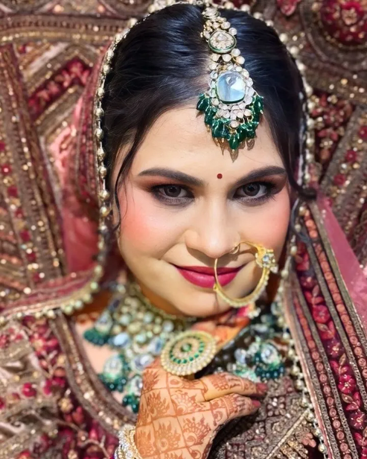 A close portrait of the same bride looking at camera, showing smoky eye makeup, a large gold nath and a green kundan maang tikka.