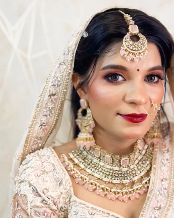 A bride in an ivory and blush lehenga with a matching veil looks at camera wearing layered kundan and pearl necklaces, a pink maang tikka and a nath.