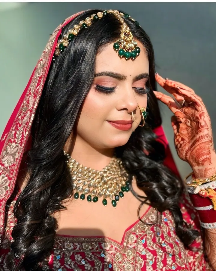 A bride in a red sequinned dupatta looks down with one hennaed hand at her temple, wearing a kundan choker with green drops and a matha patti.