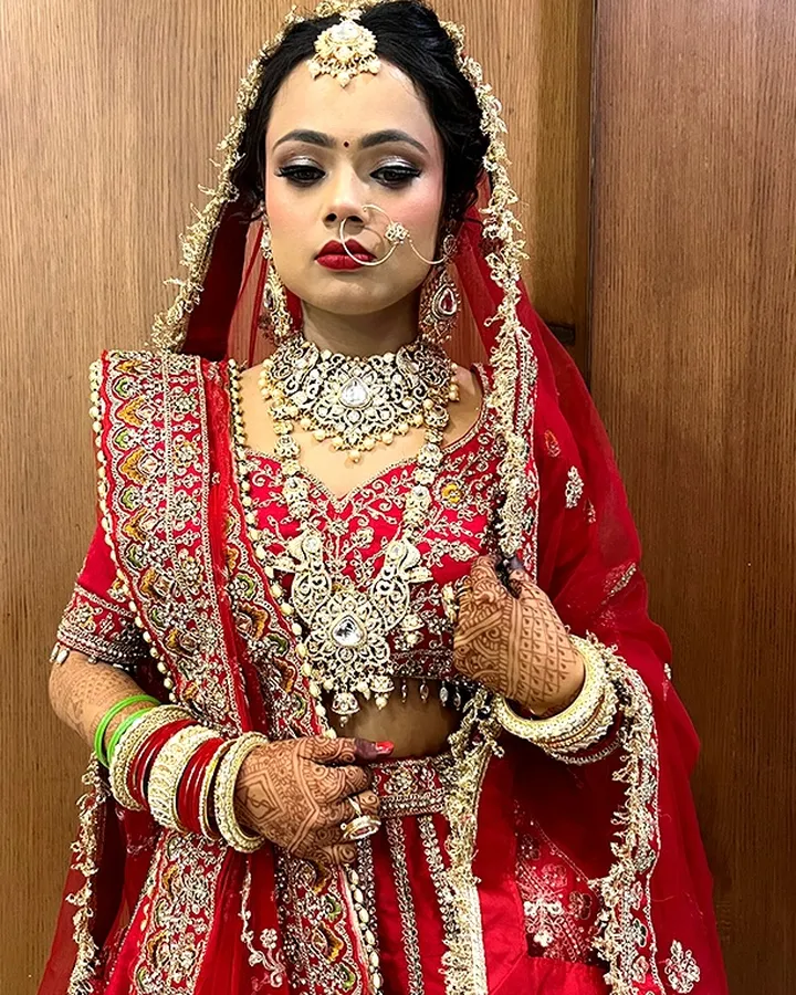 A bride in a red and gold embroidered lehenga stands against a wooden panel wearing a heavy kundan rani haar, a nath with a chain and red and green bangles.