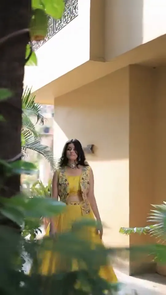 Haldi look from the styled shoot: a yellow mirror-work jacket and skirt walking past potted palms.
