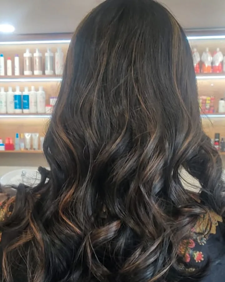 Back view of long dark hair curled at the ends, warm caramel ribbons running through it, in front of a lit salon product shelf.