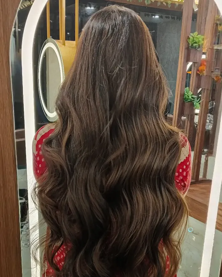 A woman photographed from behind in front of an arched, LED-lit mirror, her waist-length wavy hair carrying warm brown highlights.