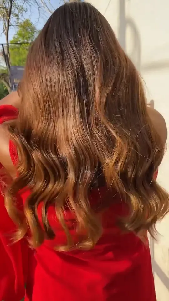 Back view of a woman in a red sleeveless outfit standing outdoors in late-afternoon sun against a white wall, long hair falling to mid-back in loose waves with .