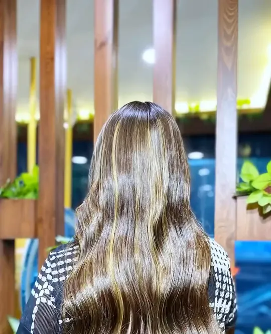 A woman photographed from behind in a black-and-white printed top, showing waist-length wavy hair in a warm brown base with clearly defined caramel and blonde h.