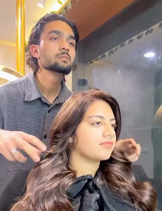 A young woman with long, heavily curled caramel-and-brown hair sits looking off to the right; a male stylist in a grey shirt stands behind her with one hand nea.