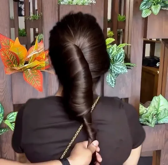 Back view of long layered hair in a warm toasted brown, lifted and let fall against a wood-slat salon wall hung with plants.