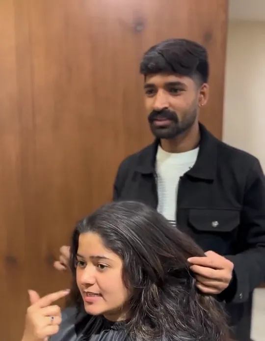 A male stylist in a black jacket stands behind a seated female client in a black cape, lifting and sectioning her hair while she talks and points at it.