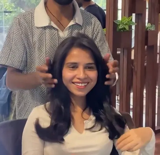 A woman client sits in a styling chair in a white long-sleeve top, smiling straight at the camera with freshly blow-dried, bouncy mid-length hair; she lifts one.