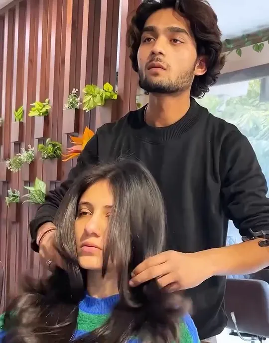 A young woman sits smiling in a black professional cape with her hair wet and combed forward while a stylist in a black sweatshirt works on it, a comb in one ha.