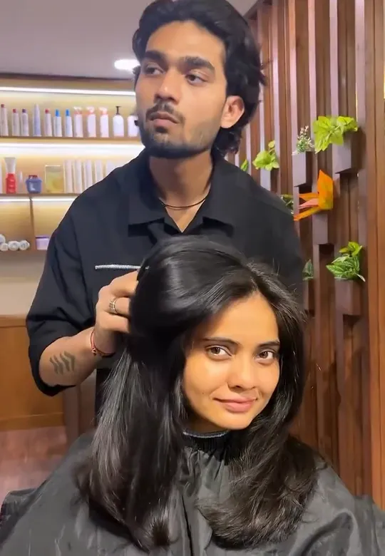 A stylist in black stands behind a seated woman and lifts her freshly cut, blow-dried shoulder-length hair with both hands.