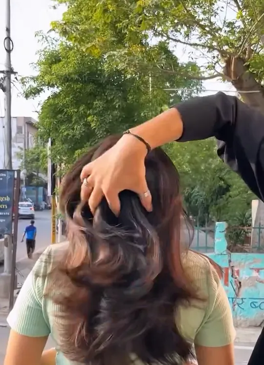Shot outdoors on a street — trees, parked cars, a metal railing and a Hindi-painted wall behind.