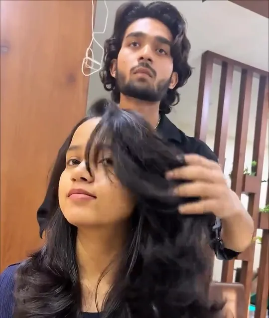 A woman sits in a salon chair while a stylist standing behind her lifts and fans out her freshly blow-dried, layered hair.