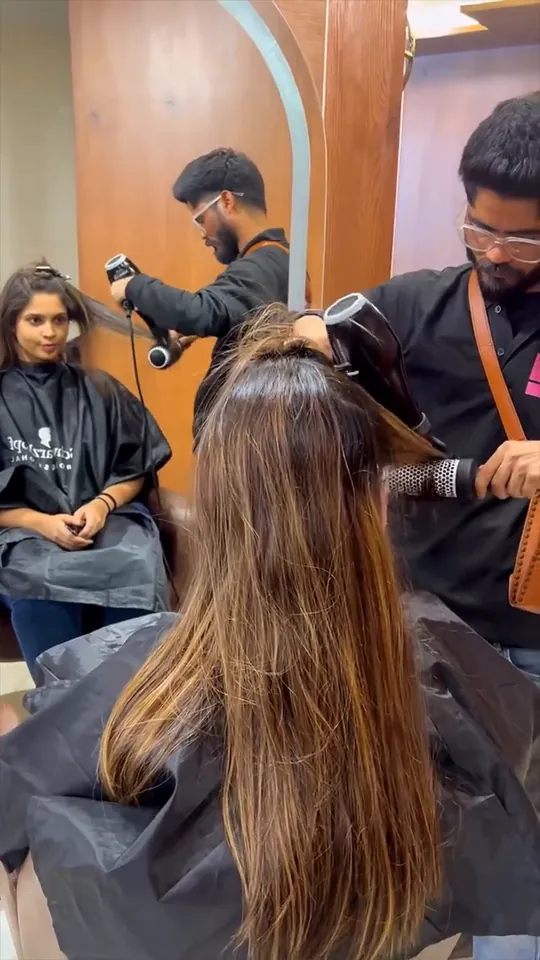 Inside the salon: a bearded male stylist in a black Glam Studios polo with a tan crossbody bag lowers a silver hood dryer over a seated female client.