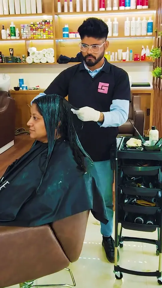A stylist in gloves paints smoothening cream through a seated client's sectioned hair, then blow-dries it out.