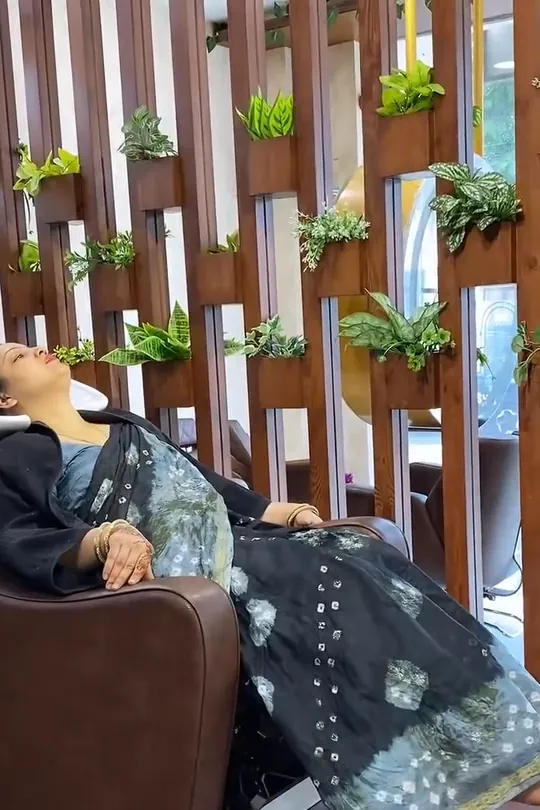 A woman in a blue-grey printed saree sits in the salon with long, poker-straight glossy black hair falling well past her waist; she wears a red lip and a bindi .