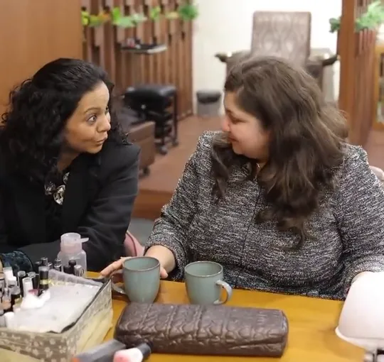 One locked-off wide shot, held for the whole reel: two women sit side by side at the nail counter with two grey-green mugs in front of them, laughing and turnin.