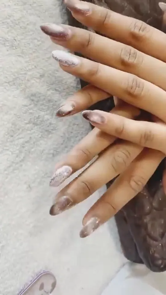 Overhead close-up of two hands crossed over each other on a white towel, showing ten long almond-shaped nail extensions in a nude-champagne glitter and cat-eye .