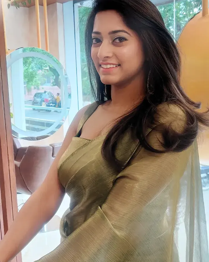 A woman in an olive-gold tissue saree turns over her shoulder to smile, standing by the salon's glass front.