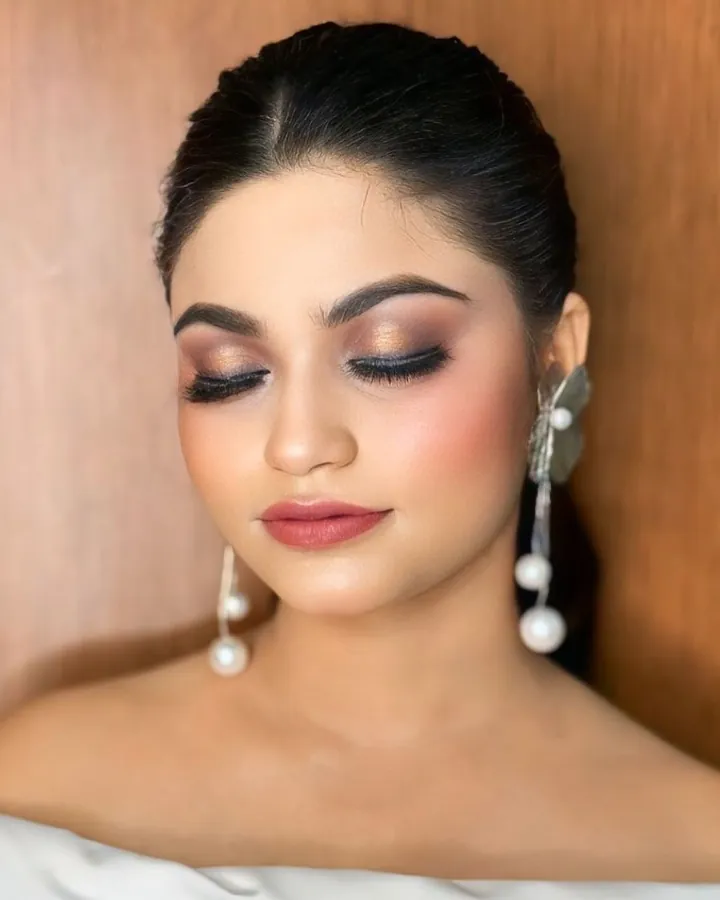 A front portrait of a woman with her eyes closed, showing bronze smoky eye makeup and a matte berry lip.