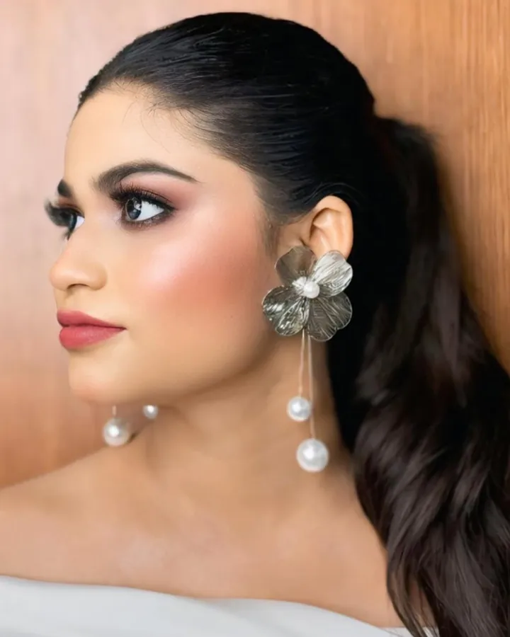 A profile portrait of a woman with a sleek high ponytail, bronze eye makeup and a silver flower drop earring.