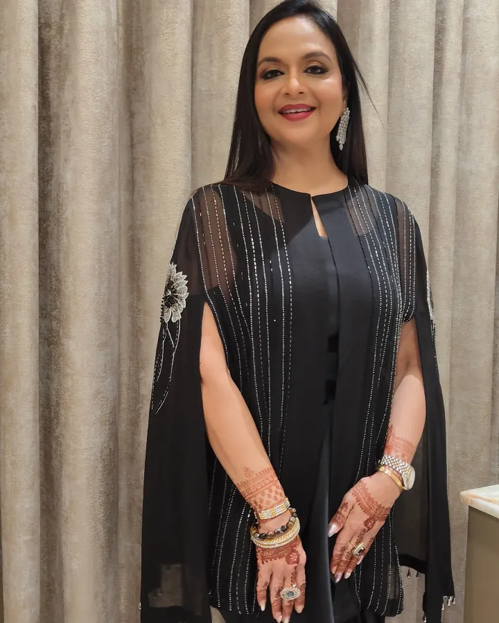 A woman in her forties or fifties stands against a champagne velvet curtain in a black cape-sleeve outfit with silver thread stripes and beaded floral motifs on.