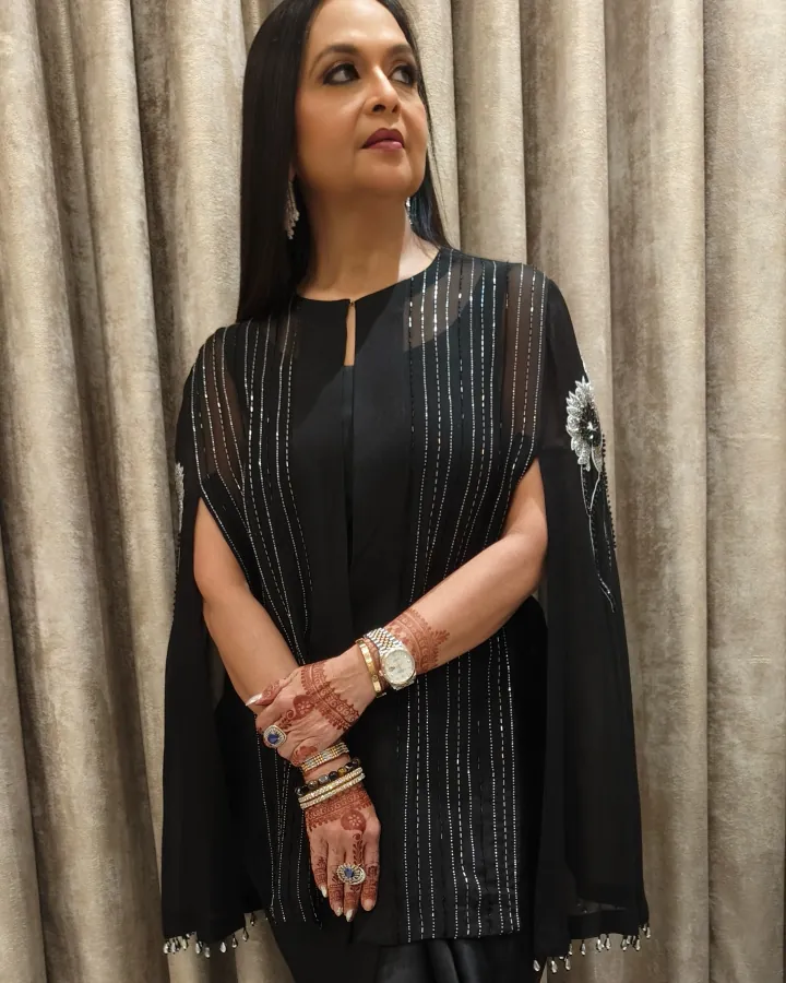 A woman in her forties or fifties stands against a champagne velvet curtain in a black cape-sleeve outfit with silver thread stripes and beaded floral motifs on.