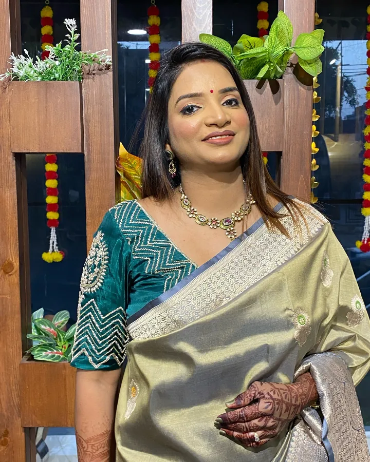 A woman in a gold tissue saree with a teal embroidered blouse and a kundan necklace stands inside the salon in front of the wooden slat partition, which is hung.