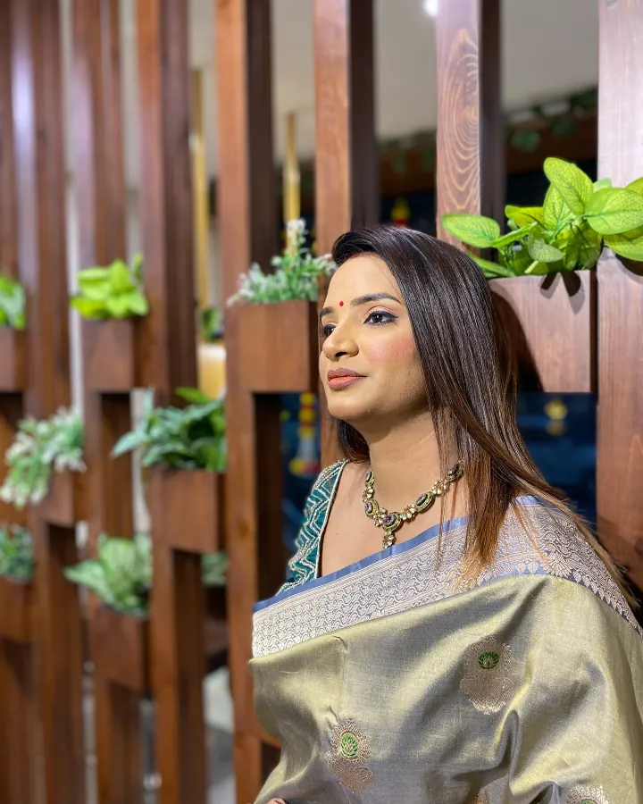 A woman in a gold tissue saree with a teal embroidered blouse and a kundan necklace stands inside the salon in front of the wooden slat partition, which is hung.