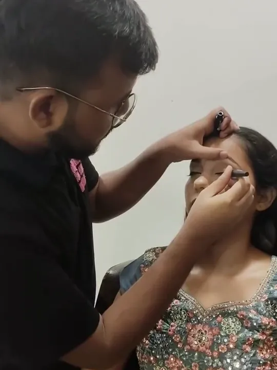 A stylist applies eye makeup with a brush to a seated client in an embroidered outfit.