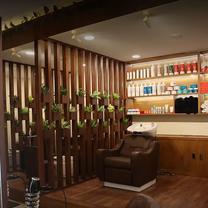 A brown recliner at a white shampoo basin beside a wooden slat partition hung with planters, with lit shelves of hair products and folded towels along the wall behind.