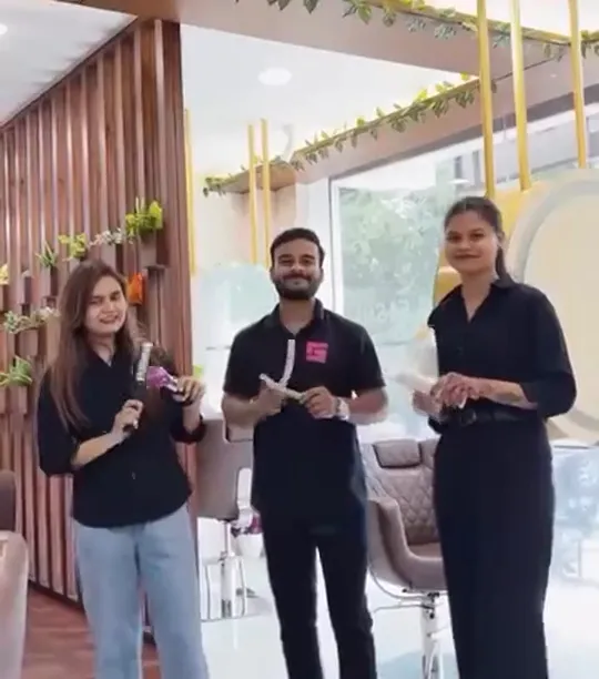 Three team members in black uniforms stand in a row on the salon floor holding styling tools, with a wooden slat partition on one side and a bright window behind them.